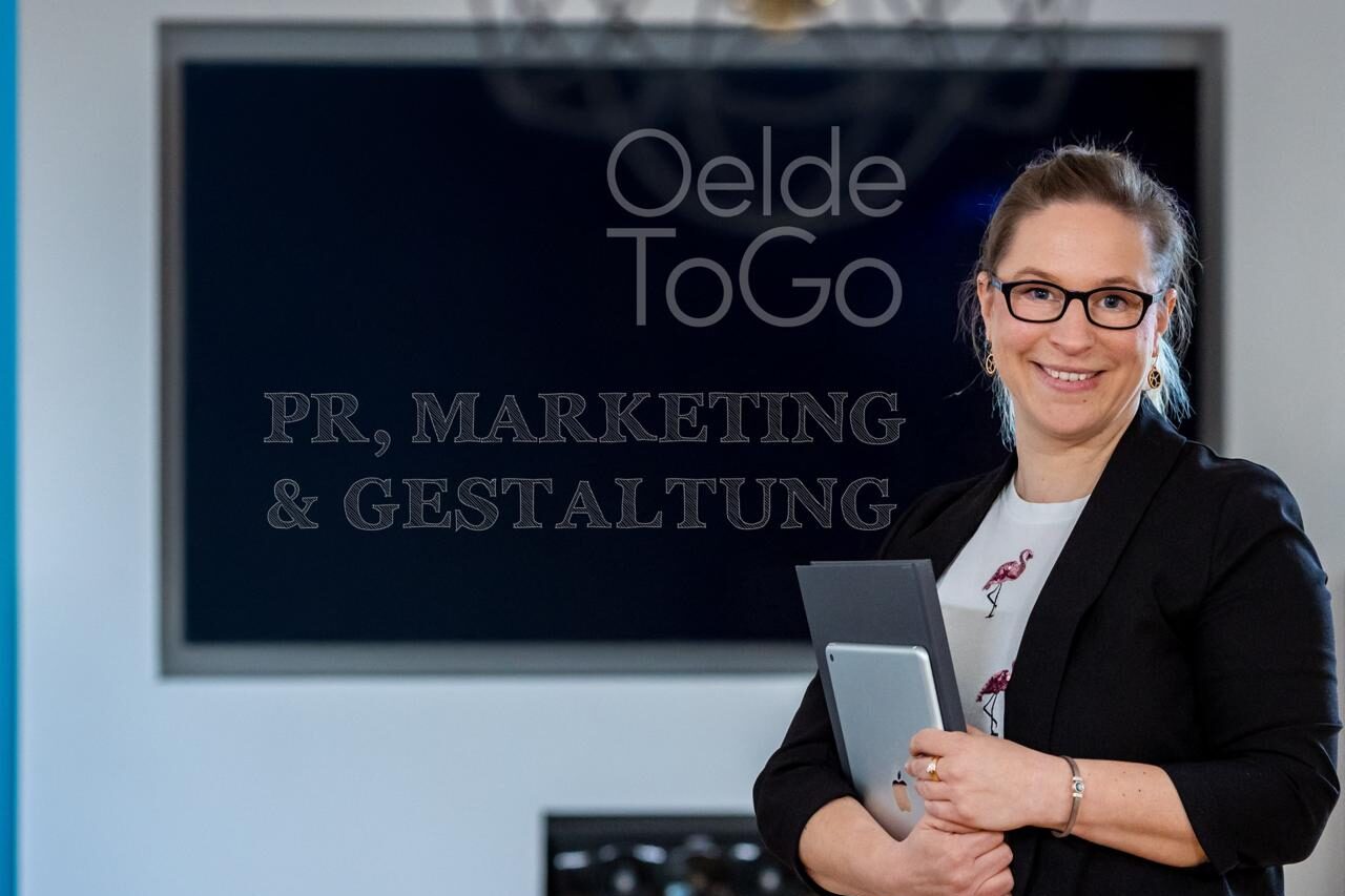 Linda Adolf, Herausgerberin der Stadtapp OeldeToGo und verantwortlich für Konzept, Design und Podcast, lächelnd mit Tablet vor dem Logo von OeldeToGo.
