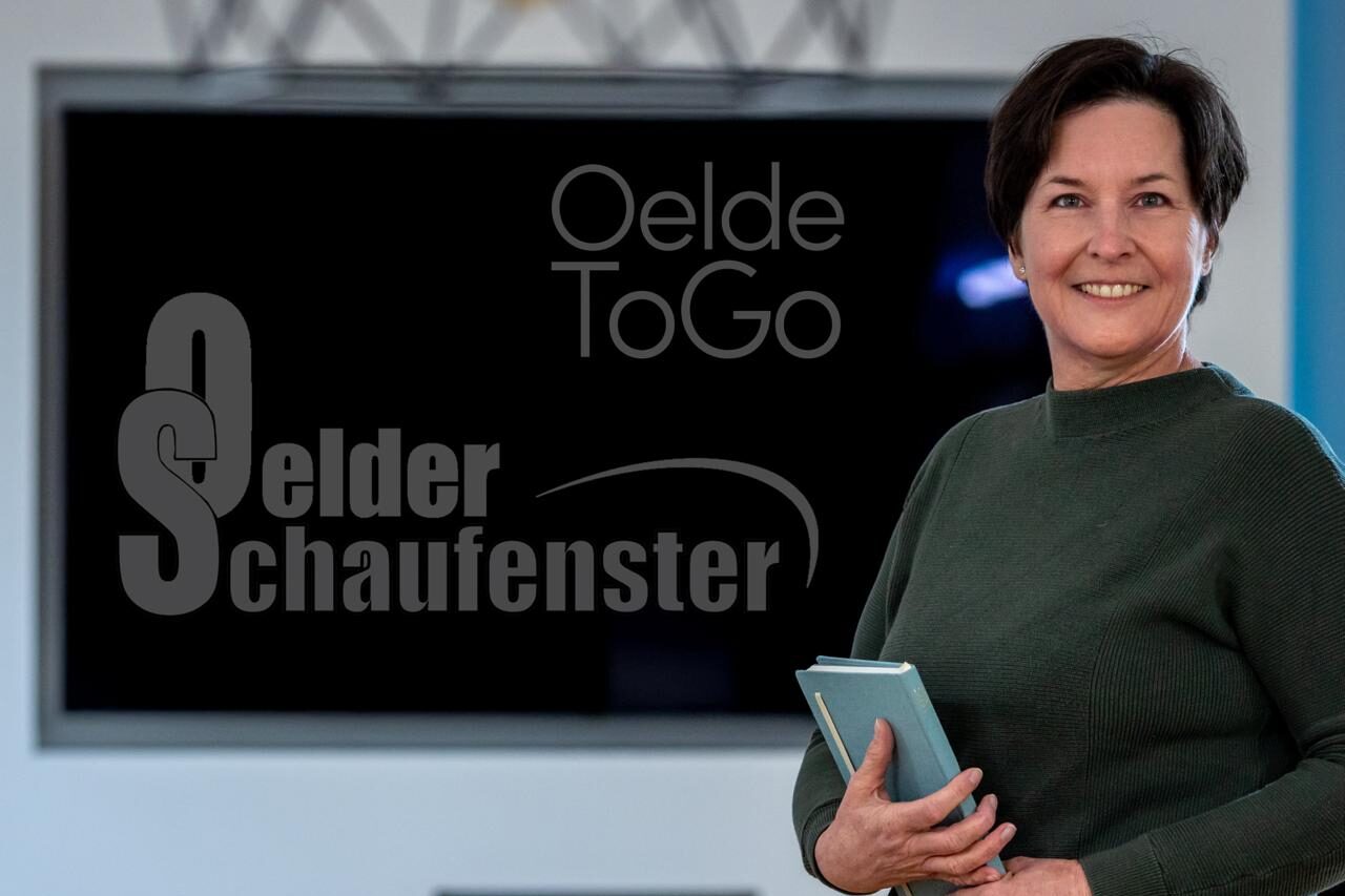 Sylvia Nübel, Herausgeberin des Oelder Schaufensters, lächelnd mit Tablet vor dem Logo von OeldeToGo und Oelder Schaufenster.